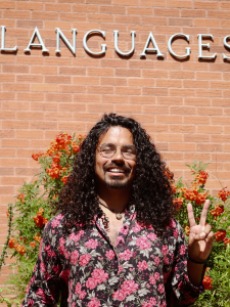 A picture of a queer black male with long curly hair and a flowers stamped blouse smiling with a hand a pace and love sign. Background bricks, orange flowers and languages sign.